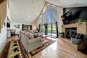 A spacious living room with a high ceiling, a fireplace, a television, and a large window overlooking a deck.