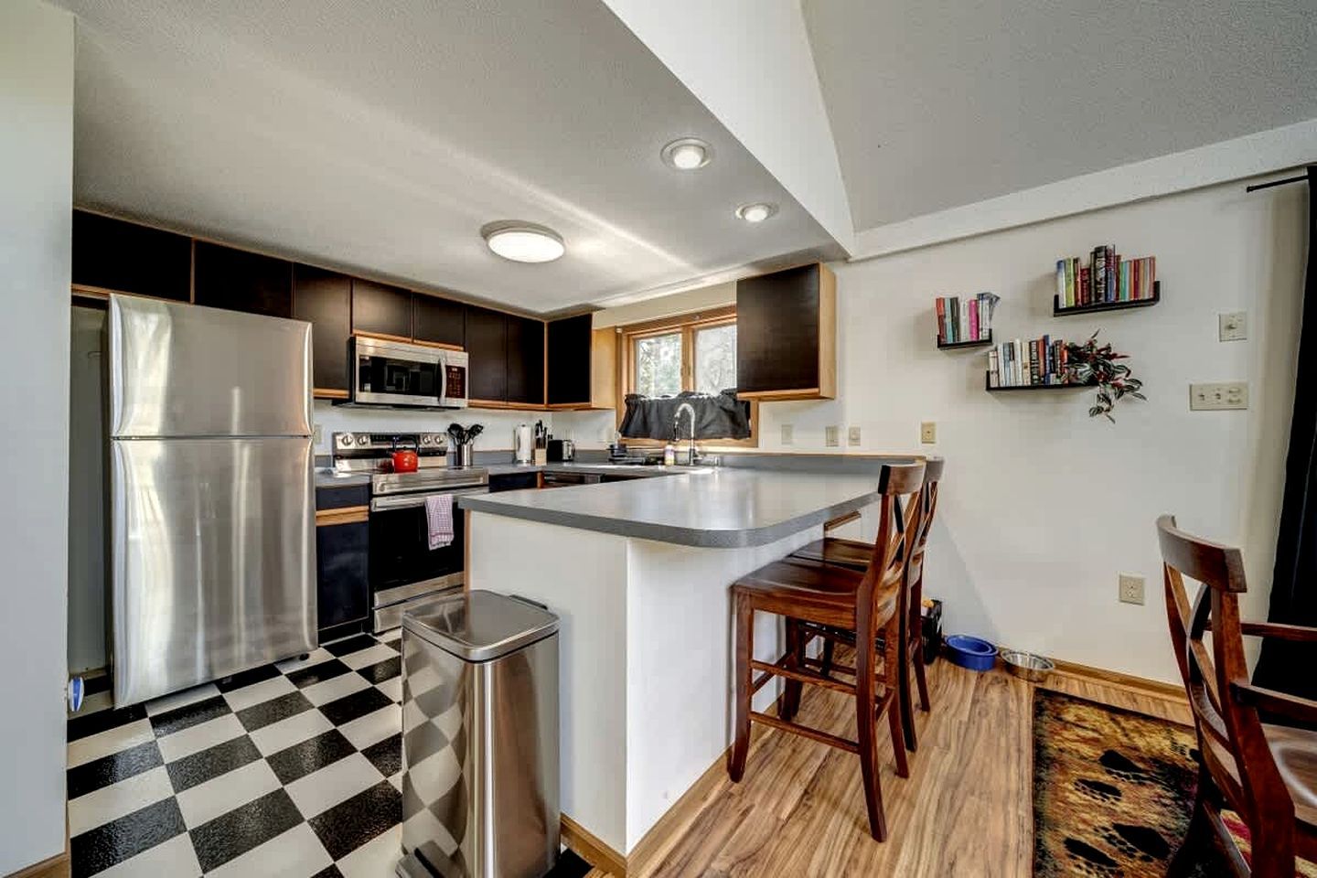 A modern kitchen with a stainless steel refrigerator, microwave, and oven, a checkered floor, and a small dining area with a wooden table and chairs.