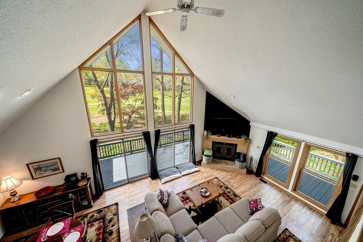 A spacious loft with a high ceiling, large windows offering a view of trees, a fireplace, and a cozy seating area.