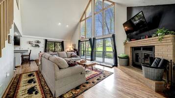 A spacious living room with a high ceiling, a fireplace, and a large window offering a view of the outdoors.