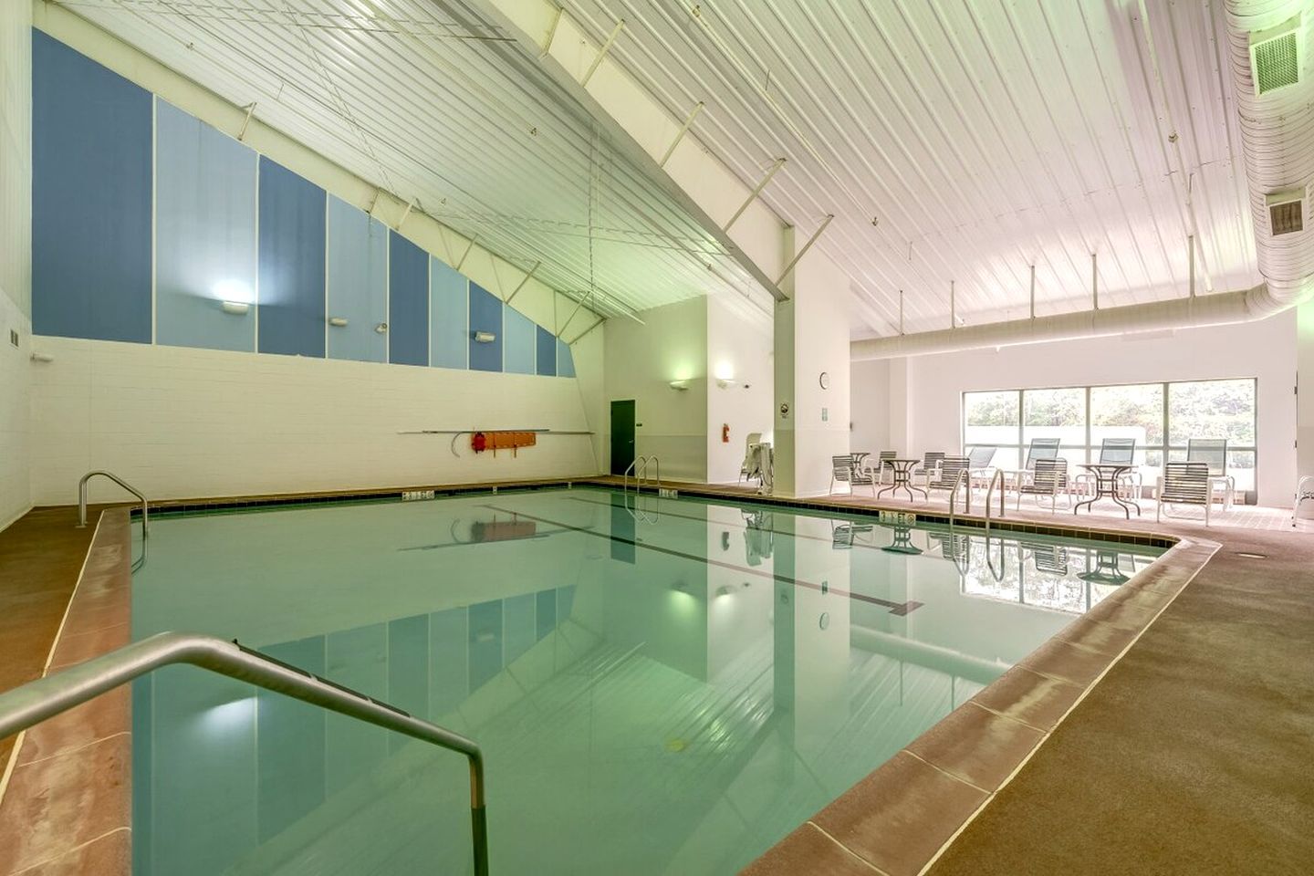 An indoor swimming pool with a tiled floor, a metal pool ladder, and a seating area with chairs and tables.