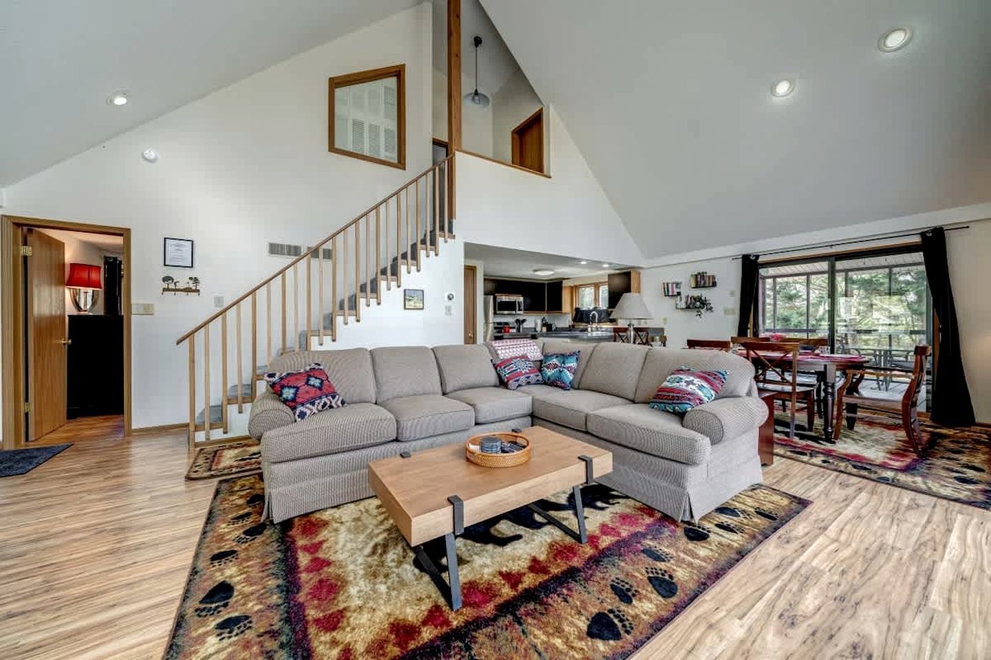 A spacious living room with a large sectional sofa, a wooden coffee table, and a patterned rug. A staircase leads to an upper level, and a dining area is visible in the background.