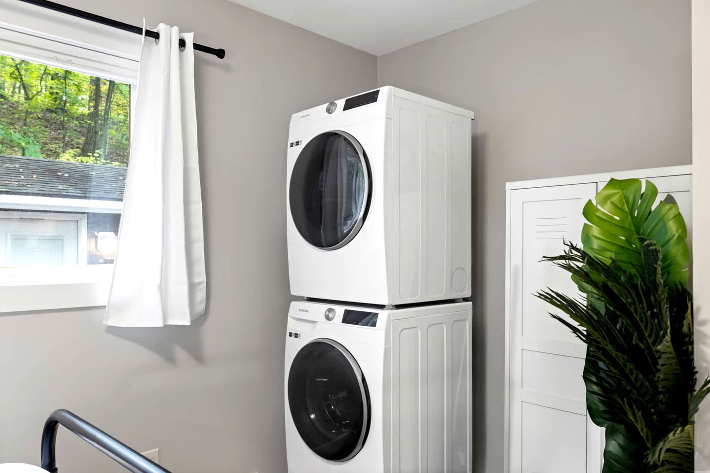 A laundry room with two stacked washing machines, a white curtain, a potted plant, and a door.