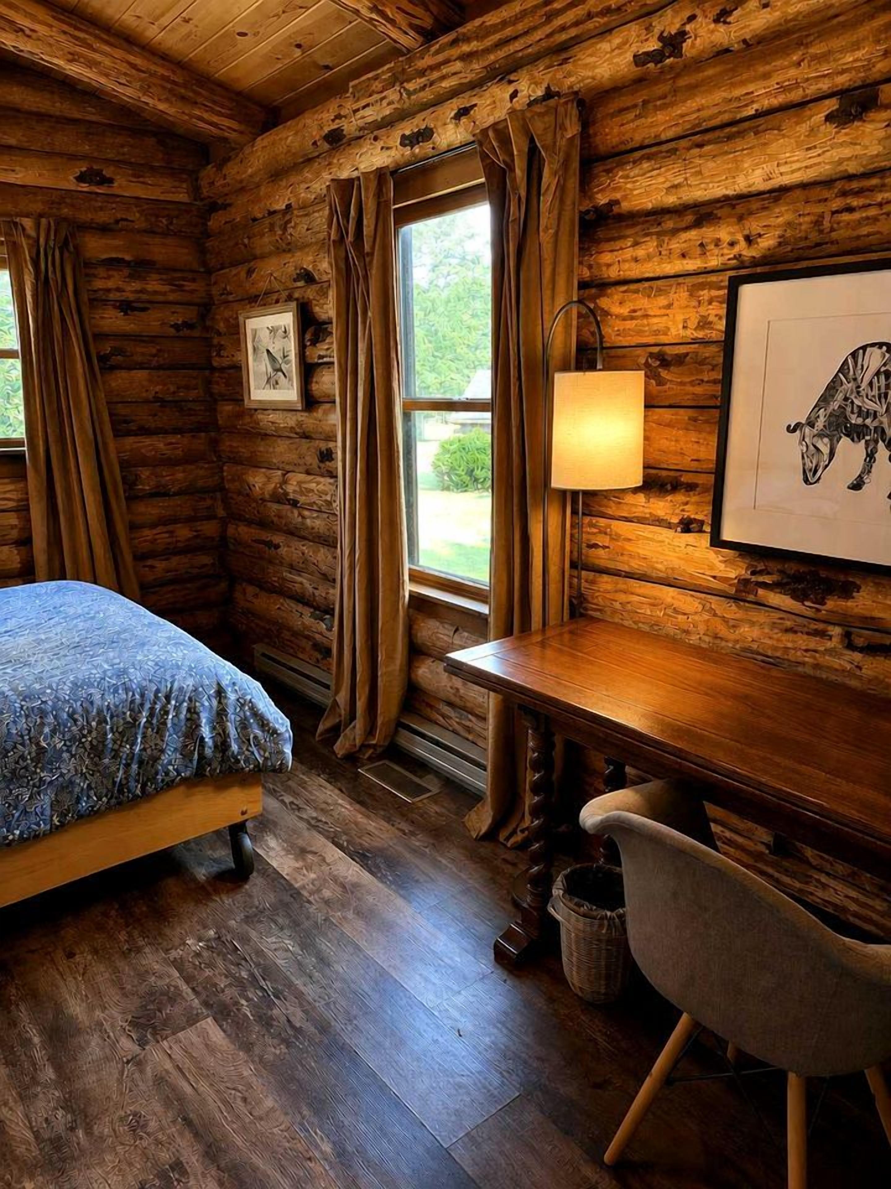 Habitación tipo cabaña de madera con cama, escritorio, silla y ventana con cortinas.