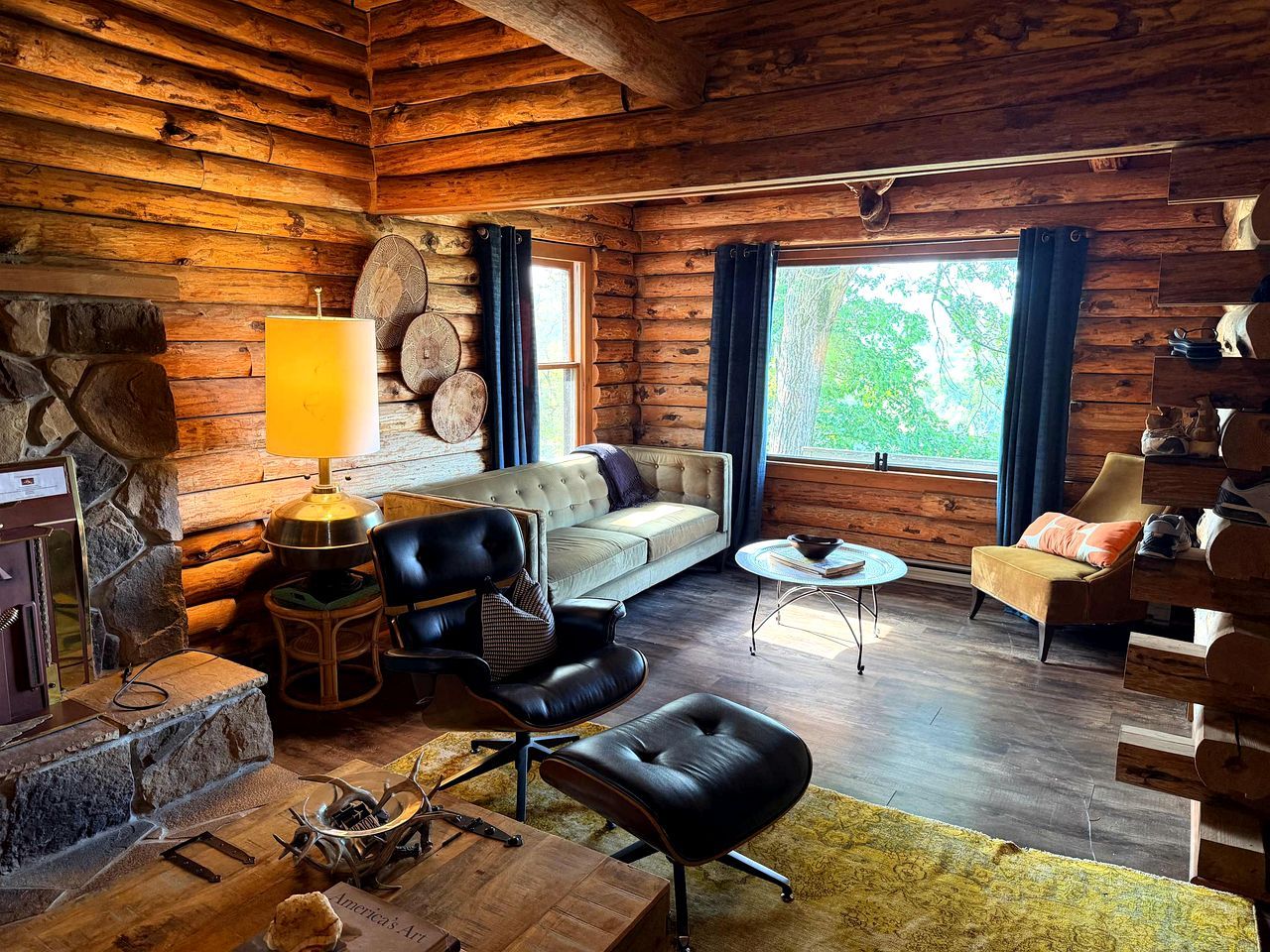 L’intérieur d’une cabane en rondins, avec une cheminée en pierre, des meubles en cuir et une grande fenêtre agrémentée de rideaux.