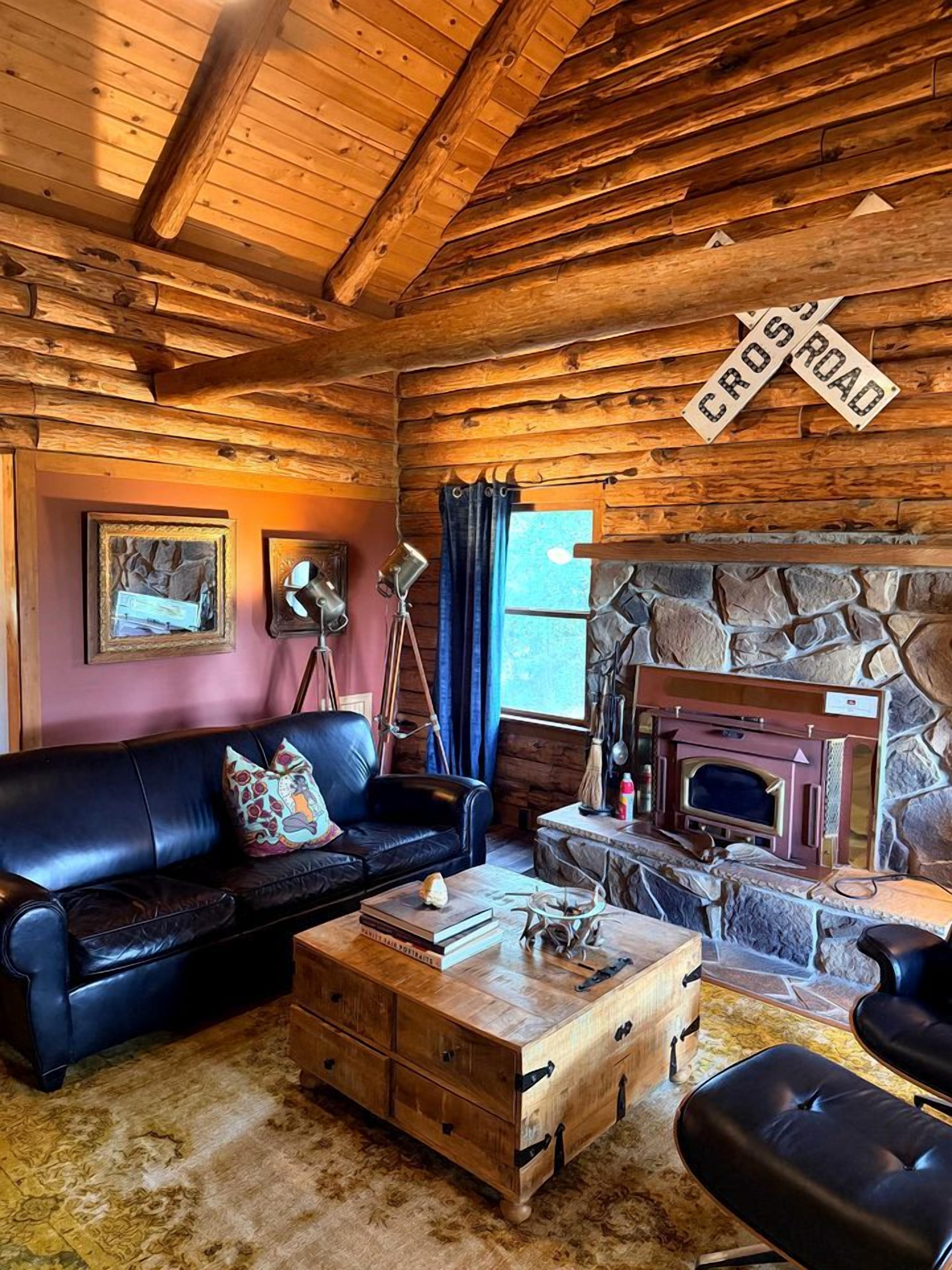 Interior rústico de una cabaña con techo de madera, chimenea de piedra, sofá de cuero y una lámpara antigua.