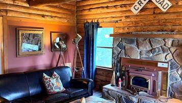 Interior rústico de una cabaña con techo de madera, chimenea de piedra, sofá de cuero y una lámpara antigua.