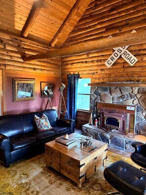 A rustic cabin interior with a wooden ceiling, a stone fireplace, a leather sofa, and a vintage lamp.
