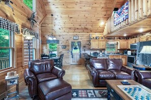 A cabin interior with leather sofas, a wooden ceiling, a kitchen area, and a view through a door. - Mountain View Cabin w/ Hot Tub & Game Room (Mineral Bluff)