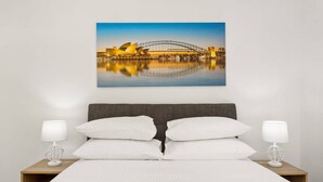 A hotel room with a bed, two bedside lamps, and a wall-mounted picture of the Sydney Opera House.