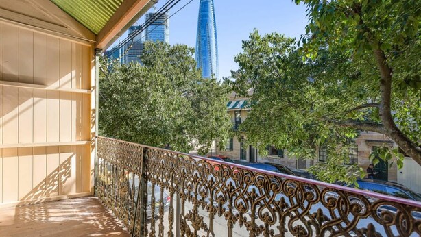 A balcony with ornate metal railings, a wooden floor, and a view of a cityscape with tall buildings and trees.