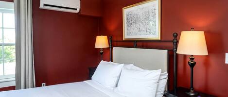 A hotel room with a bed, two bedside lamps, an air conditioning unit, a framed artwork on the wall, and a window with curtains.