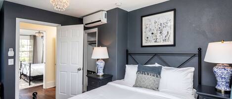 A bedroom with a bed, bedside lamps, a nightstand, a dresser, a chandelier, an air conditioning unit, a door to another room, and a framed artwork on the wall.