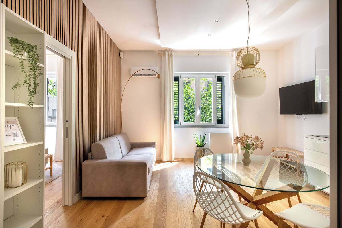Un salon moderne avec une table à manger en verre, des chaises blanches, un canapé beige et un téléviseur fixé au mur.