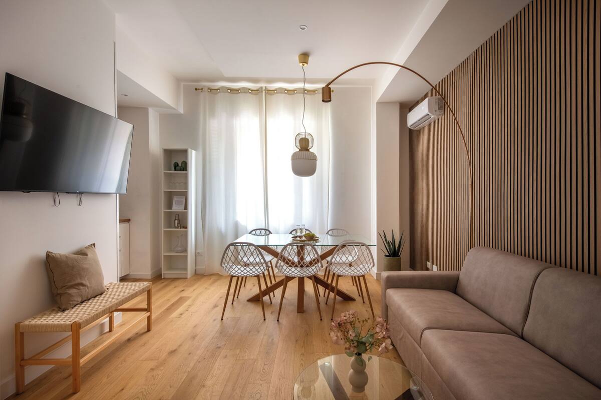 Un salon moderne comprenant un canapé, un banc, une table à manger avec des chaises, un téléviseur fixé au mur et un luminaire suspendu.
