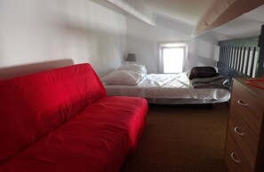 Ein Zimmer mit einem roten Sofa, einem Bett mit weißer Bettwäsche, einem hölzernen Schminktisch und einem Fenster mit Jalousien.