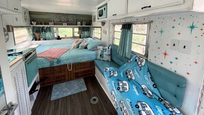 A compact RV bedroom with a bed, a small kitchenette, and a window with curtains.