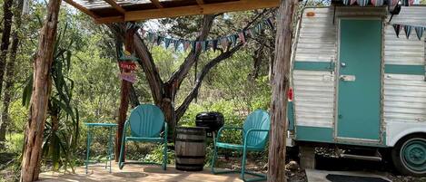 A covered outdoor seating area with a 'WELCOME' sign, a vintage camper, and a tree.