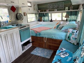 Ein kompaktes Wohnmobil-Interieur mit einer Küchenzeile, einem Bett mit vielen Kissen und einem Fenster mit Vorhängen.