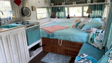A compact RV interior with a kitchenette, a bed with multiple pillows, and a window with curtains.