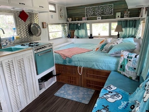 A compact RV interior with a kitchenette, a bed with multiple pillows, and a window with curtains.