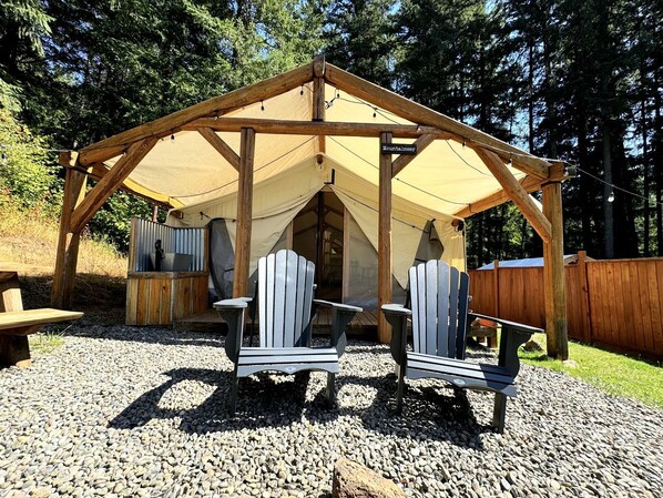 A wooden gazebo with a tent, two Adirondack chairs, and a grill.