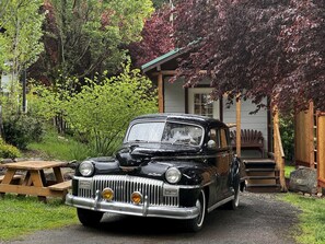 Un coche negro antiguo estacionado frente a una casa con terraza de madera y escalones que llevan a un porche.