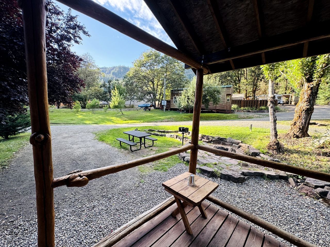 A wooden deck with a picnic table and benches, overlooking a park with a lake and mountains.