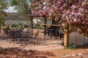 An outdoor patio with tables and chairs, a pergola with pink flowers, and a building in the background.