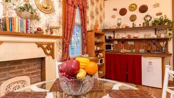 A dining area with a table set for two, a fruit bowl, a fireplace, and a kitchen area with shelves and appliances.
