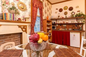 A dining area with a table set for two, a fruit bowl, a fireplace, and a kitchen area with shelves and appliances.
