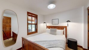 Ein Schlafzimmer mit einem Holzbett, einem Fenster mit Jalousien, einem Spiegel, einer Nachttischlampe und einem gerahmten Bild an der Wand.