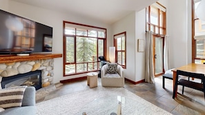 A living room with a stone fireplace, large windows, a flat-screen TV, a sofa, a chair, and a dining table.