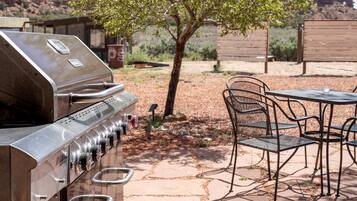 A stainless steel grill, outdoor seating with chairs and a table, and a tree in a landscaped area.