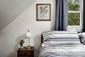 A bedroom with a bed, a nightstand, a lamp, a framed botanical illustration, and a window with curtains.