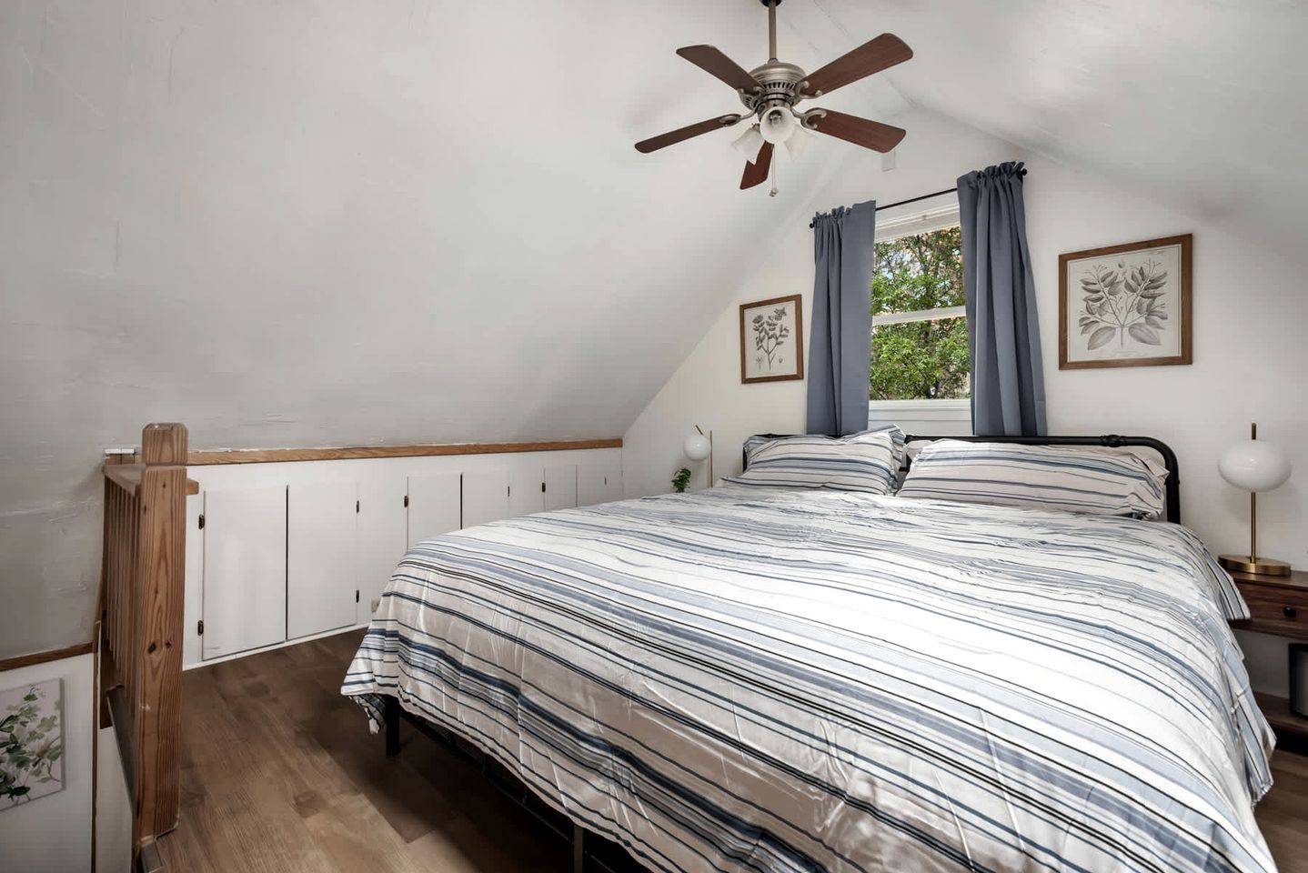 Quarto com teto inclinado, cama com roupa de cama listrada, ventilador de teto e duas gravuras botânicas emolduradas na parede.