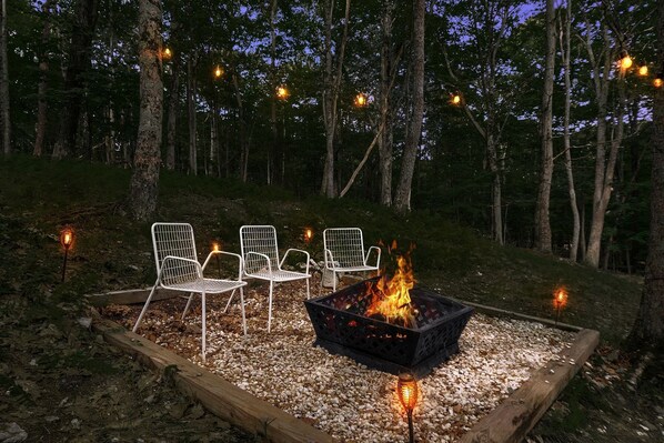 A fire pit with chairs and string lights in a forest setting.