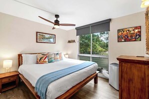 A bedroom with a bed, a ceiling fan, a window with blinds, a nightstand, a dresser, and artwork on the walls.