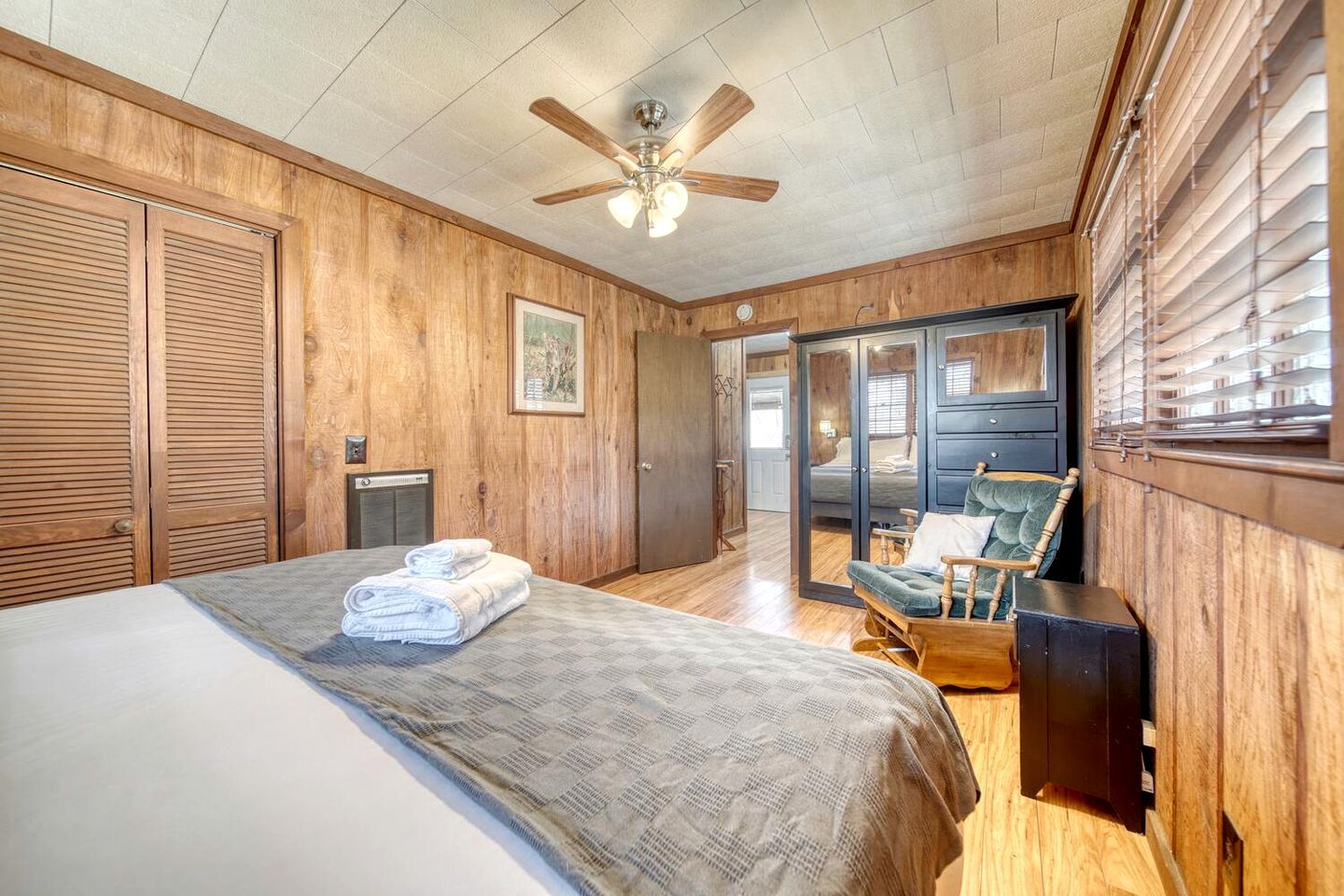 A bedroom with a bed, a wooden ceiling fan, a rocking chair, and a view through a glass door.