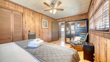A bedroom with a bed, a wooden ceiling fan, a rocking chair, and a view through a glass door.
