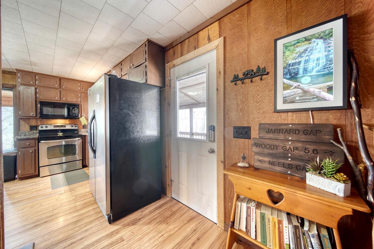 Una cocina con armarios de madera, un refrigerador negro, un microondas y un horno. Hay una estantería de madera con libros y una pequeña planta.