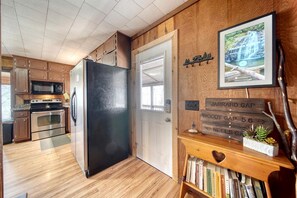 A kitchen with wooden cabinets, a black refrigerator, a microwave, and an oven. There is a wooden shelf with books and a small plant.