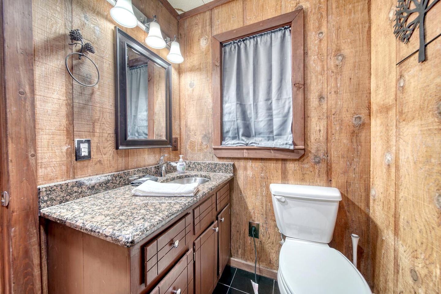 A bathroom with wooden walls, a granite countertop, a sink, a toilet, and a window with a curtain.