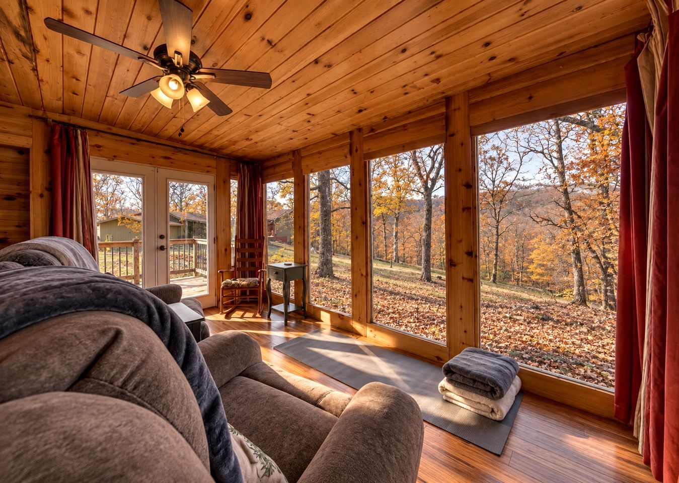 A cozy cabin interior with a wooden ceiling, a rocking chair, a small table, and a view of a forest through large windows.