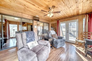 A wooden interior with a ceiling fan, a sofa, a chair, a desk, and a large window.