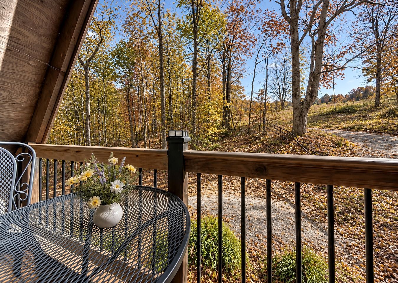 A wooden deck with a metal table and chairs, overlooking a forested area with autumn foliage.