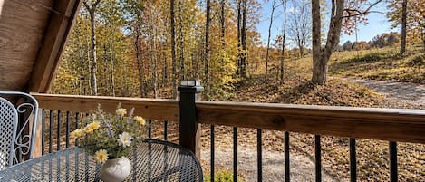 Terraza de madera con una mesa y sillas de metal, con vistas a un área boscosa con follaje otoñal.