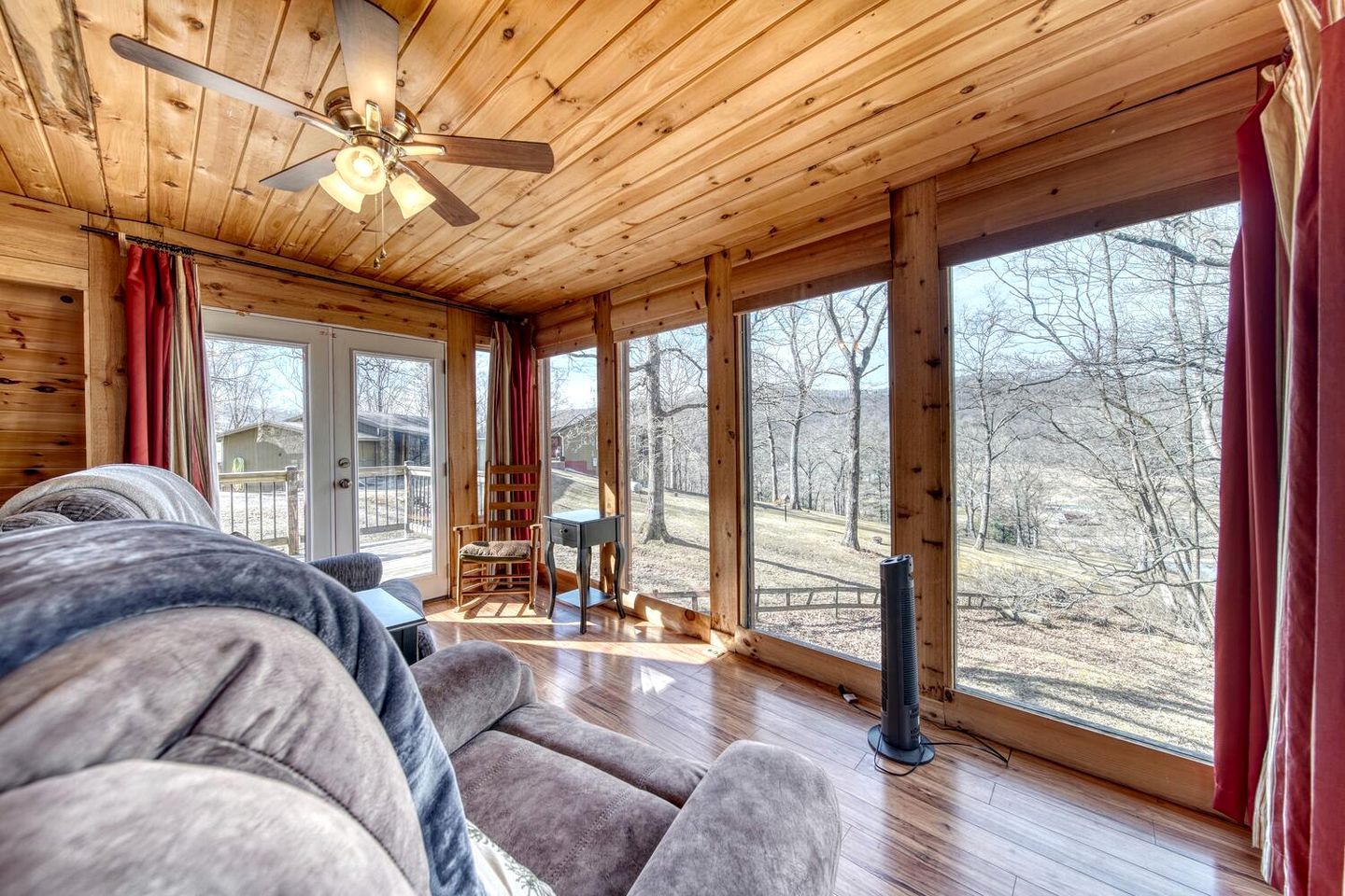 A wooden cabin interior with a ceiling fan, large windows, a sofa, a chair, and a small table.