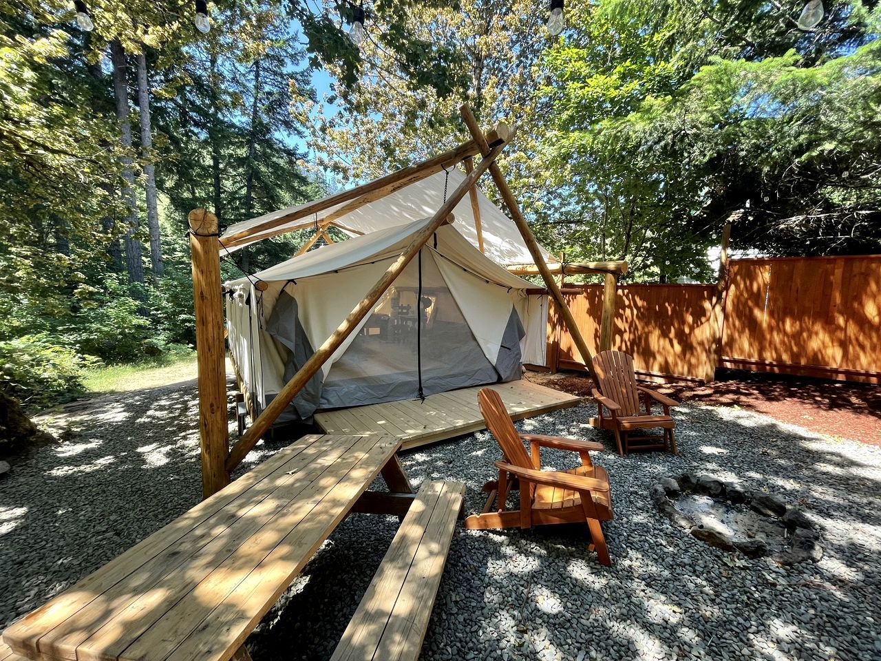 A tent with a wooden frame and a tarp roof, surrounded by a gravel area with wooden benches and a fire pit.