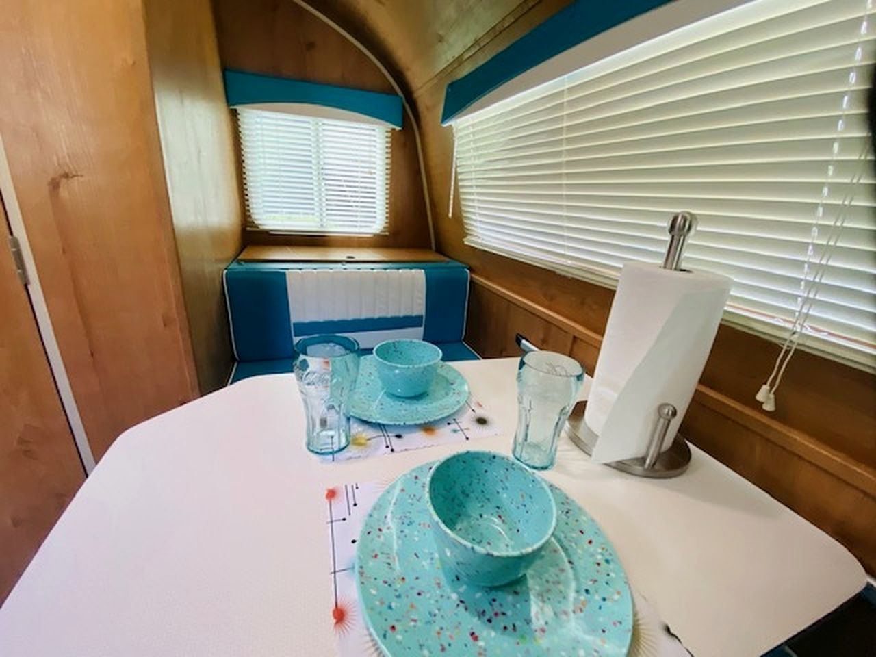Un coin repas avec une table dressée pour deux personnes, une fenêtre avec des stores et un banc à l’arrière-plan.